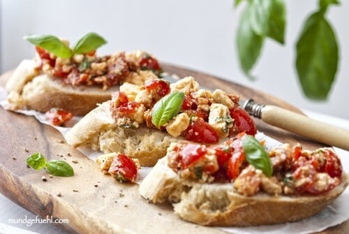 drei Stück Bruschetta mit Tomaten und Schafskäse liegen auf einem Holzbrett mit einem Messer