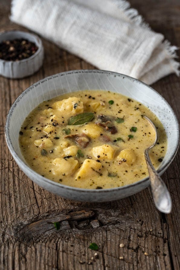 Eine Schüssel mit Kartoffelsuppe und einer Serviette auf Holzuntergrund, daneben eine Schale mit Pfefferkörnern
