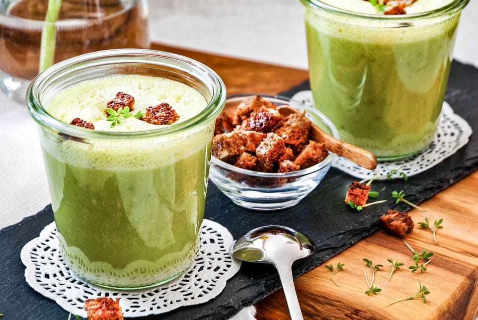 Zwei Gläser mit Erbsensuppe und einem Schälchen Croutons stehen auf einem Holzbrett