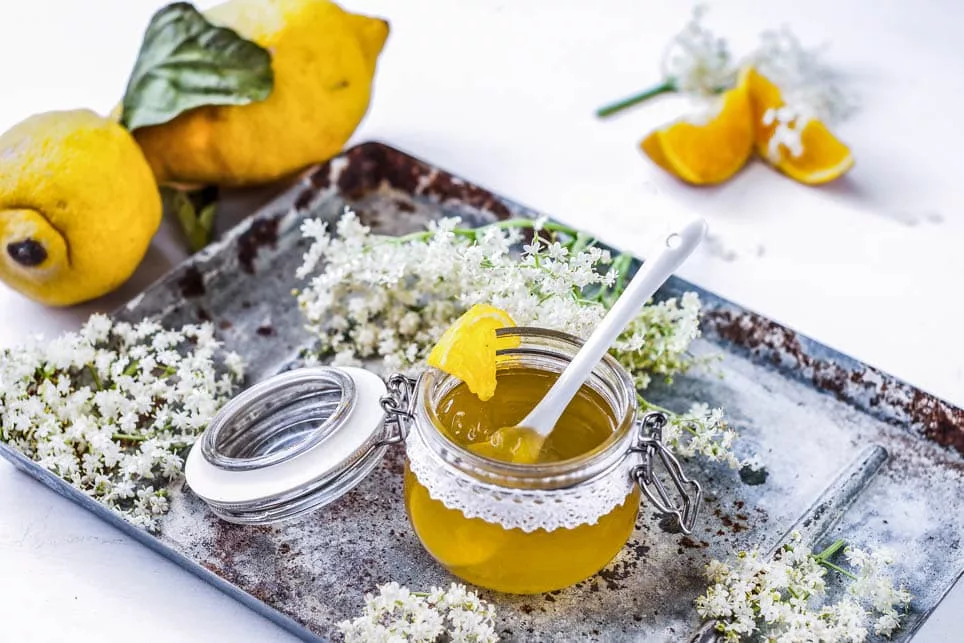 Ein Glas Holundergelee auf Tablett mit Holunderblüten daneben.