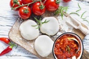 3 Kugeln Pizzateig liegen auf einem Brett neben Tomaten und einer Schüssel Tomatensauce