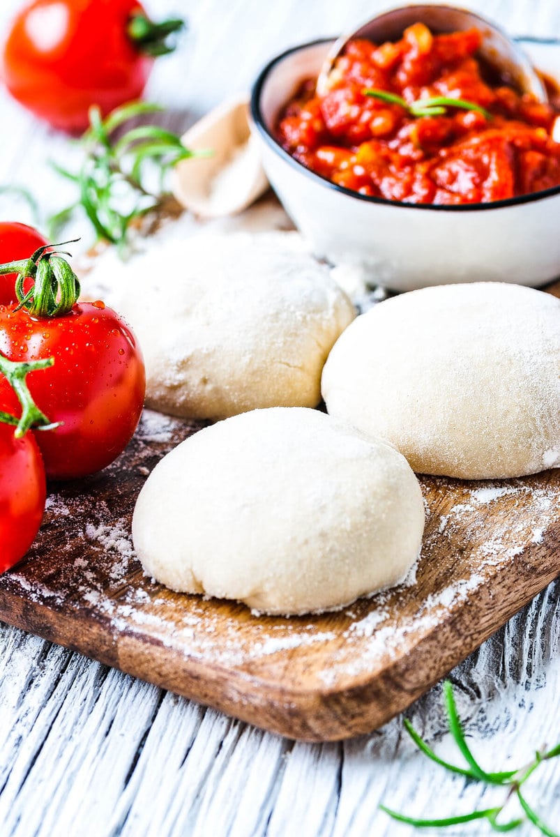 3 Kugeln Pizzateig liegen auf einem Brett neben Tomaten und einer Schüssel Tomatensauce