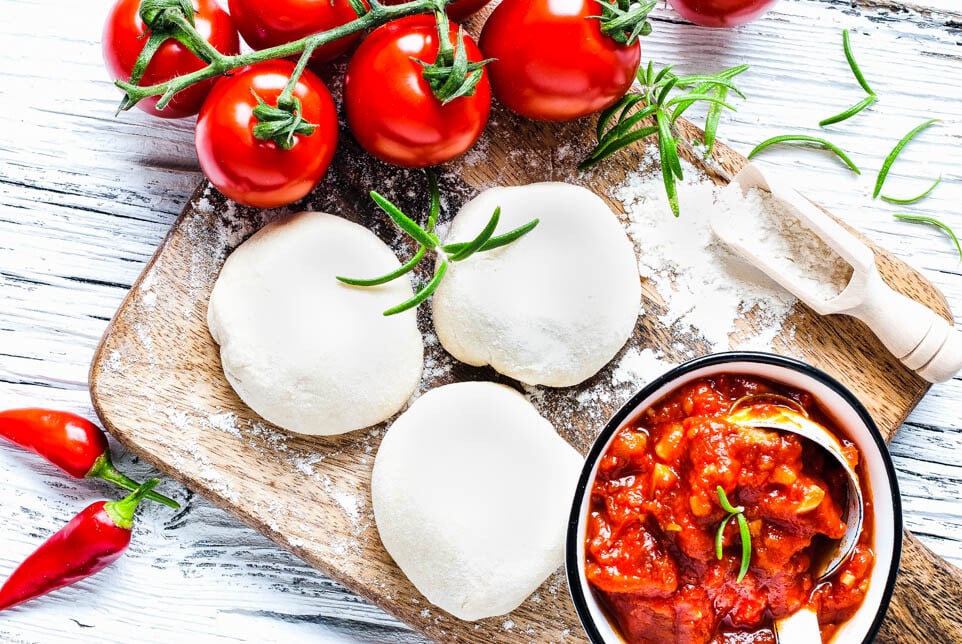 3 Kugeln Pizzateig liegen auf einem Brett neben Tomaten und einer Schüssel Tomatensauce