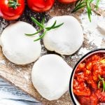 3 Kugeln Pizzateig liegen auf einem Brett neben Tomaten und einer Schüssel Tomatensauce