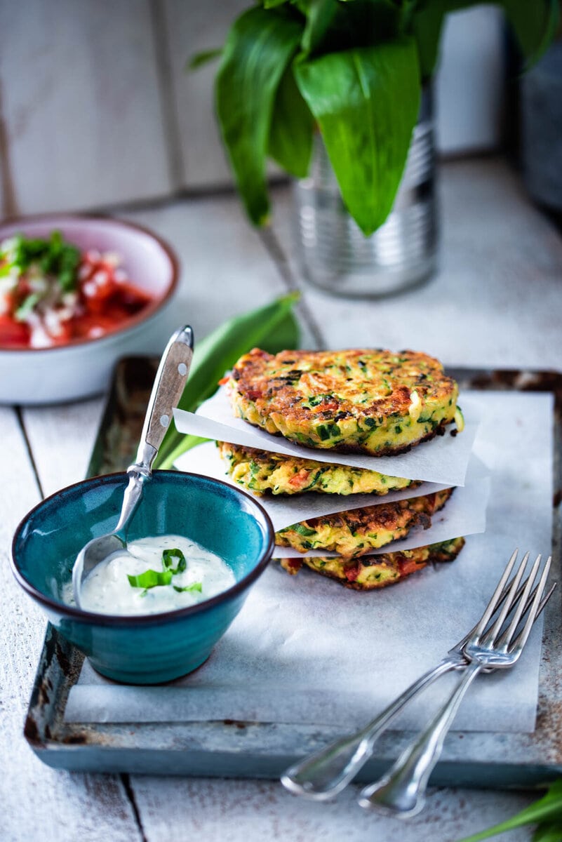 Zucchinipuffer liegen aufeinander neben einer Schüssel mit Kräutersauce und Tomatensalat