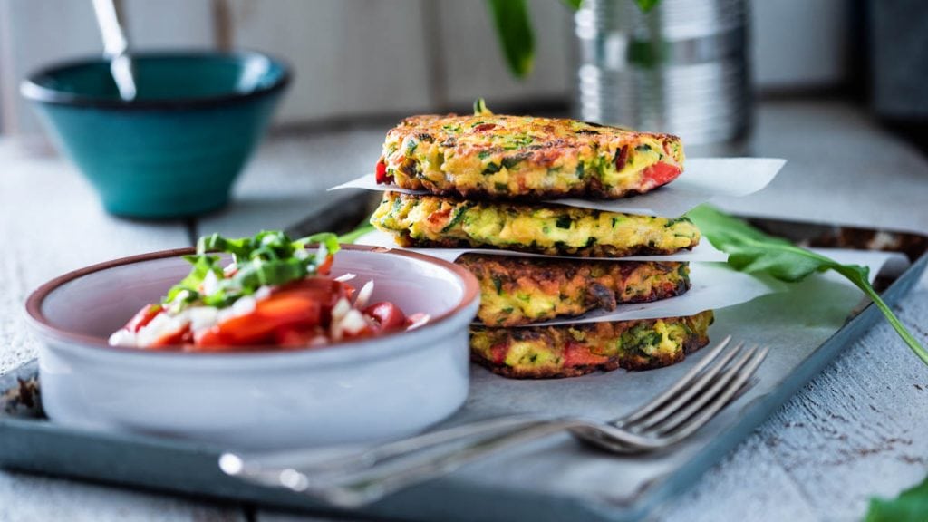 Zucchinipuffer liegen aufeinander neben einer Schüssel mit Kräutersauce und Tomatensalat