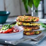 Zucchinipuffer liegen aufeinander neben einer Schüssel mit Kräutersauce und Tomatensalat