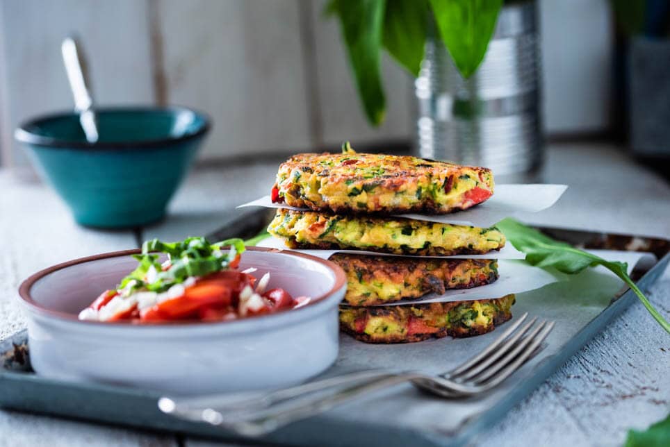 Zucchinipuffer liegen aufeinander neben einer Schüssel mit Kräutersauce und Tomatensalat