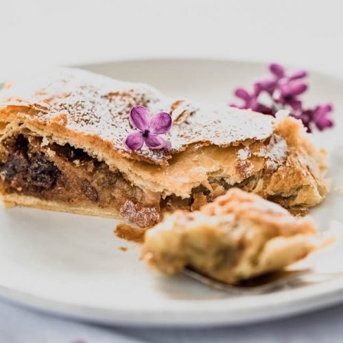 Ein Stück Rhabarberstrudel auf einem Teller, dekoriert mit Fliederblüten