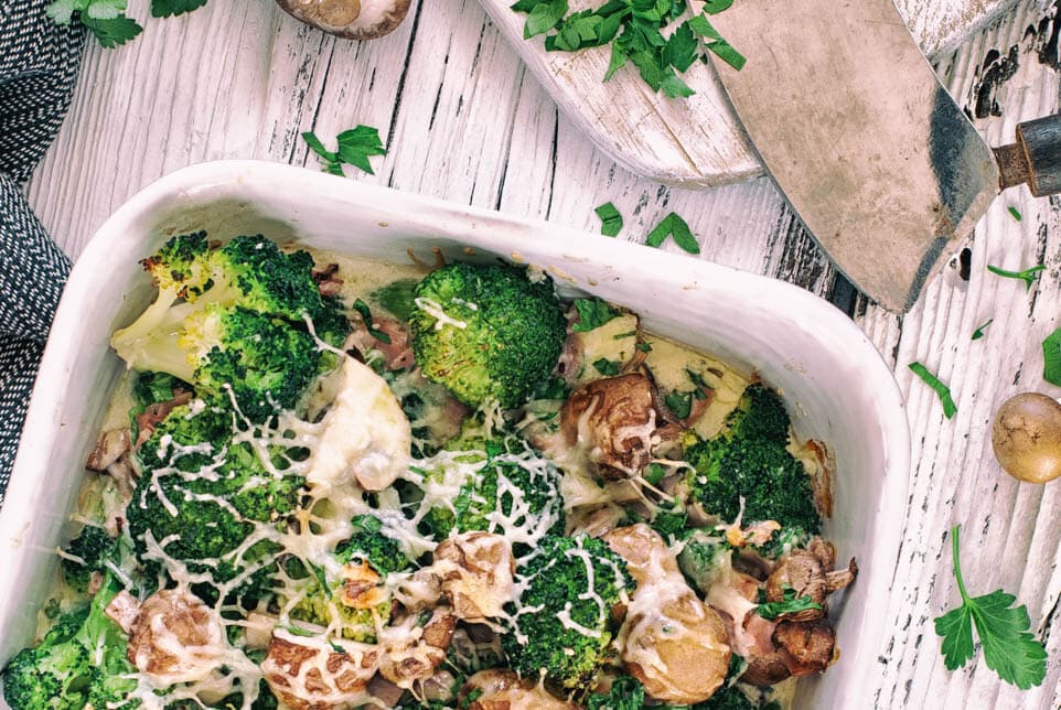 Brokkoli Auflauf mit Käse überbacken steht auf Holzuntergrund mit Champignons und Besteck
