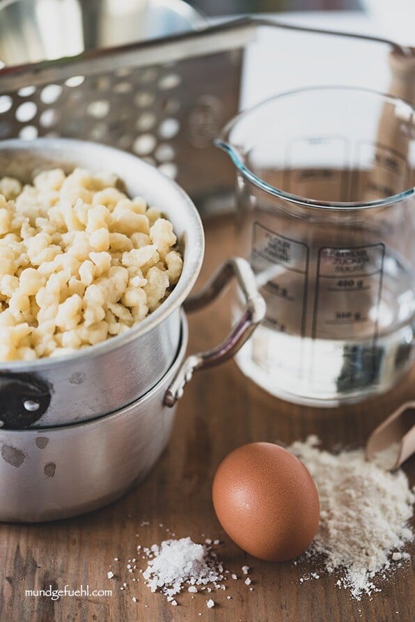 Fertige Spätzle stehen neben ihren Zutaten Mehl, Wasser, Ei und Salz