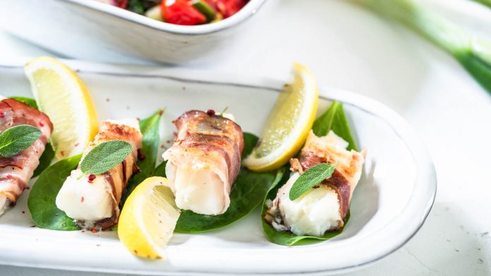 saltimbocca-vom-kabeljau-mit-mediterranem-ofengemuese Drei mit Speck umwickelte Fischstücke sind mit Gemüse und Aioli auf einem Teller angerichtet