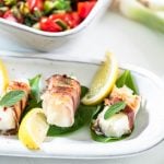 Drei mit Speck umwickelte Fischstücke sind mit Gemüse und Aioli auf einem Teller angerichtet