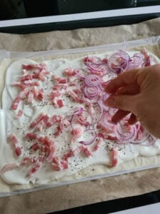 Teig wird mit Speck und Zwiebeln belegt