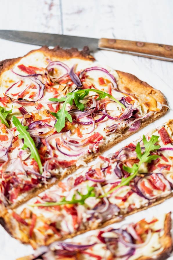 Elsässer Flammkuchen liegt aufgeschnitten auf Backpapier, daneben ein Messer.