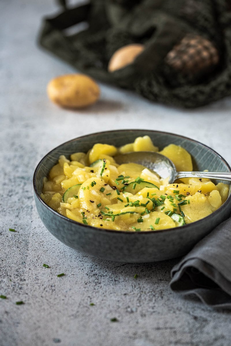 Eine Schüssel Kartoffelsalat steht auf Steinuntergrund und dahinter liegt ein Einkaufsnetz mit Kartoffeln.