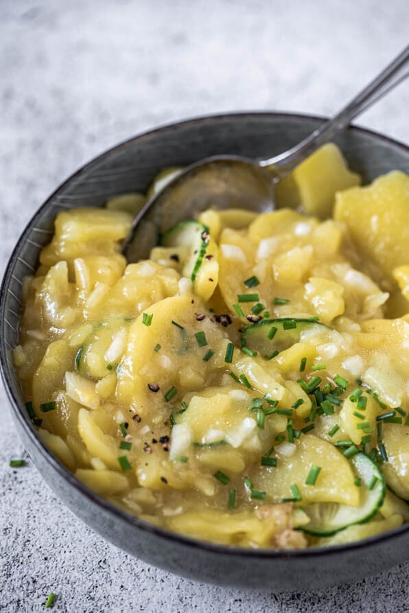 Eine Schüssel Kartoffelsalat steht auf Steinuntergrund und dahinter liegt ein Einkaufsnetz mit Kartoffeln.