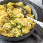 Eine Schüssel Kartoffelsalat steht auf Steinuntergrund und dahinter liegt ein Einkaufsnetz mit Kartoffeln.