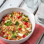 Eine Schüssel mit Couscous Salat steht mit einer Wasserflasche und einem Glas auf weißem Holzuntergrund