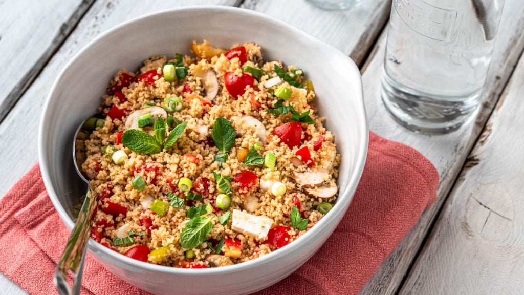 Eine Schüssel mit Couscous Salat steht mit einer Wasserflasche und einem Glas auf weißem Holzuntergrund