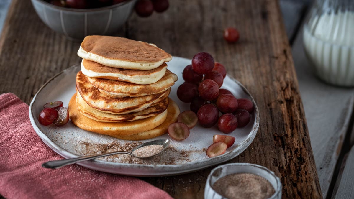 Ein Stapel Mini Pancakes mit Zimt und Zucker auf einem Teller. Dahinter Weintrauben und ein Milchkrug.