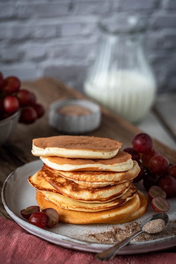 Ein Stapel Mini Pancakes ist auf einem Teller. Dahinter Milch und Trauben.