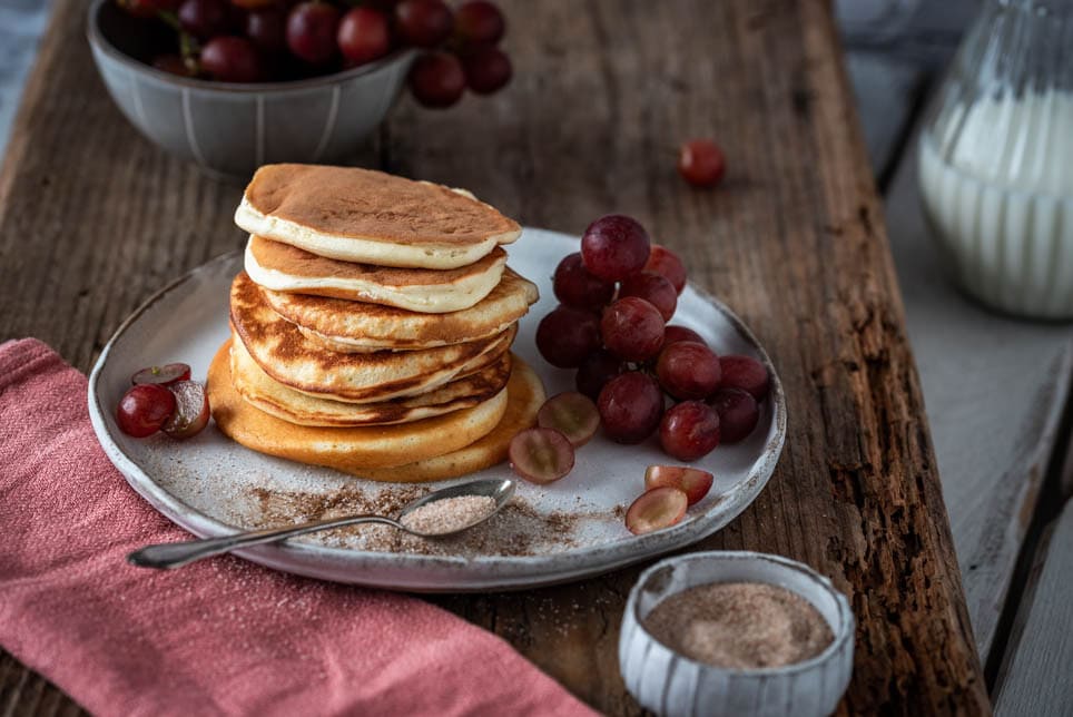 Ein Stapel Mini Pancakes mit Zimt und Zucker auf einem Teller. Dahinter Weintrauben und ein Milchkrug.