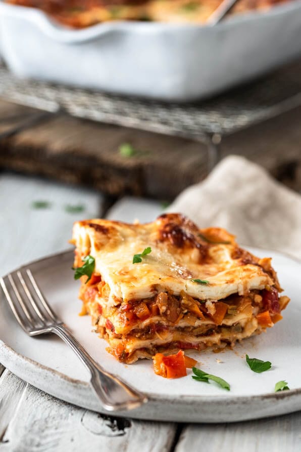 Ein Stück Gemüselasagne auf einem Teller. Im Hintergrund die Auflaufform des Ofengerichts