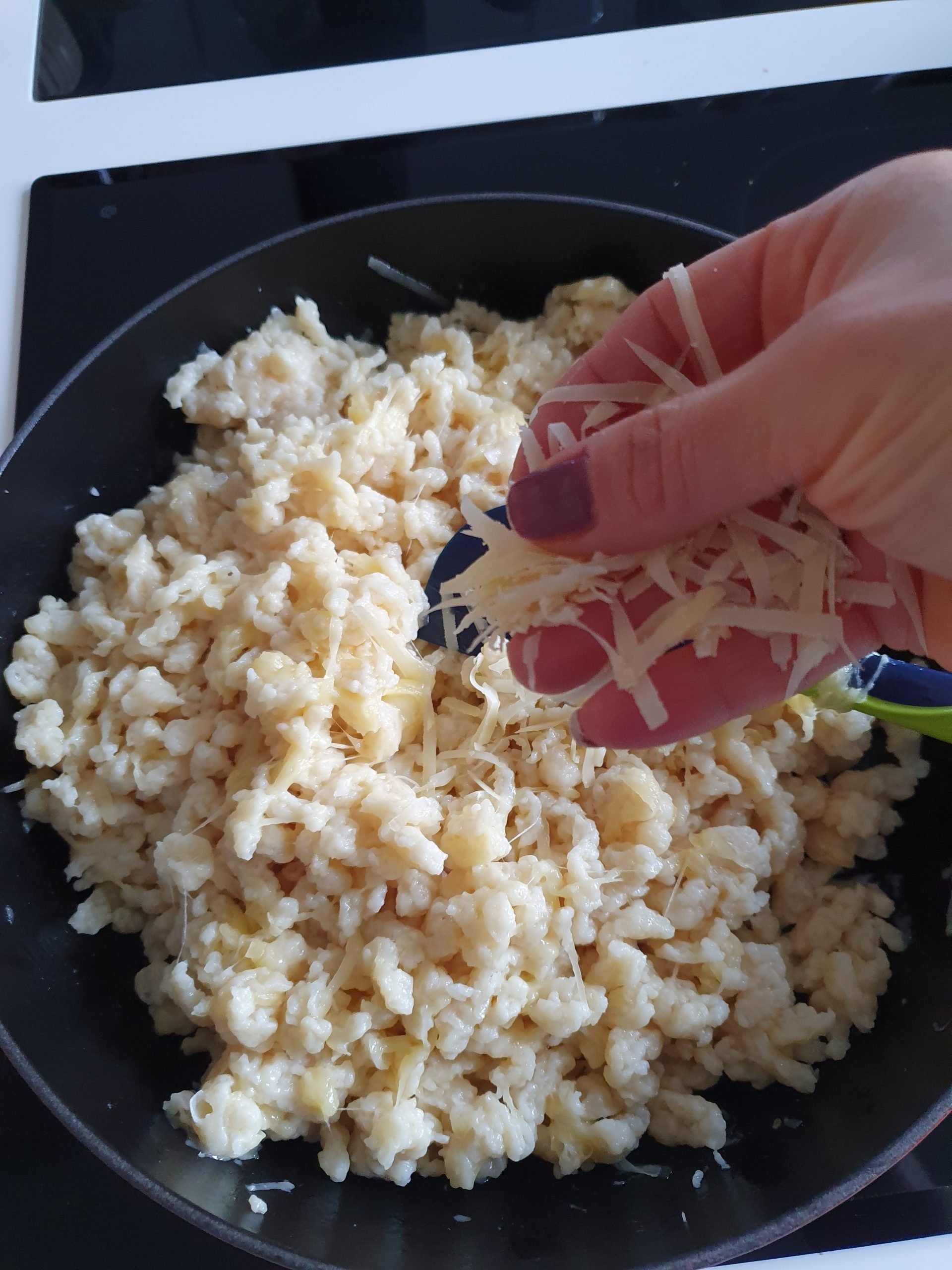 Eine Hand gibt Käse in die Pfanne mit Spätzle