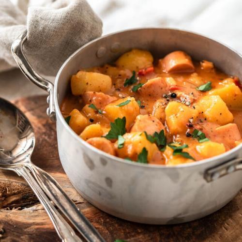 Ein Topf Kartoffelgulasch mit Würstchen steht auf einem Holzbrett.