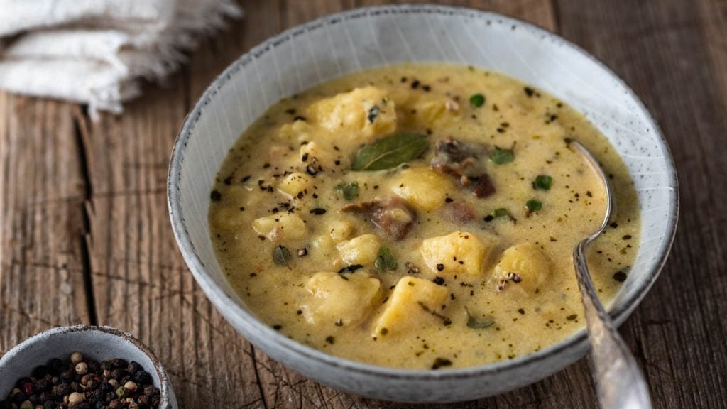 Eine Schüssel mit Kartoffelsuppe und einer Serviette auf Holzuntergrund, daneben eine Schale mit Pfefferkörnern
