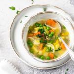 Ein Teller Gemüsesuppe in weißem Geschirr und daneben Kräuter.