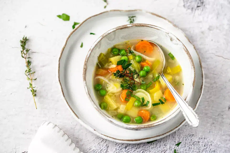 Ein Teller Gemüsesuppe in weißem Geschirr und daneben Kräuter.