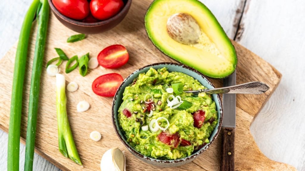 Guacamole in einer Schüssel auf Holzbrett. Daneben Avocado, Tomaten und Lauchzwiebeln