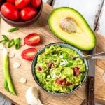 Guacamole in einer Schüssel auf Holzbrett. Daneben Avocado, Tomaten und Lauchzwiebeln