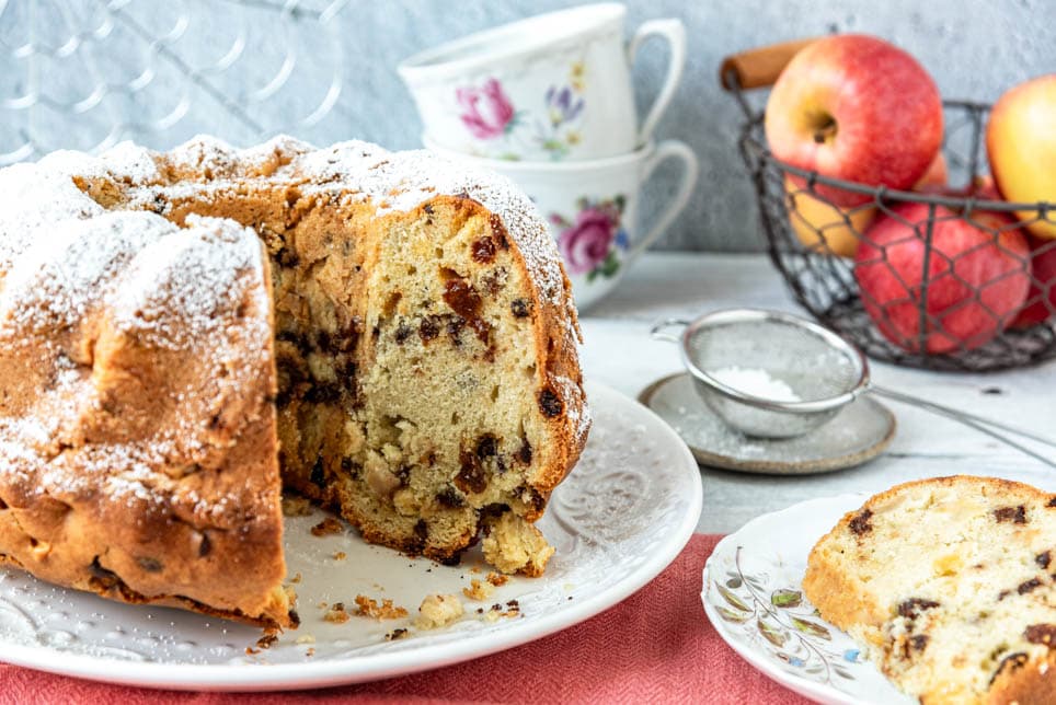 Angeschnittener Apfel-Gugelhupf mit Tassen und Äpfeln auf Tisch