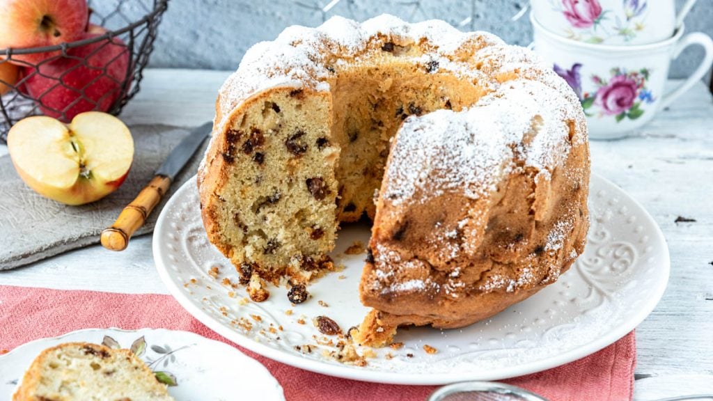 Angeschnittener Apfel-Gugelhupf mit Tassen und Äpfeln auf Tisch - Mundgefühl
