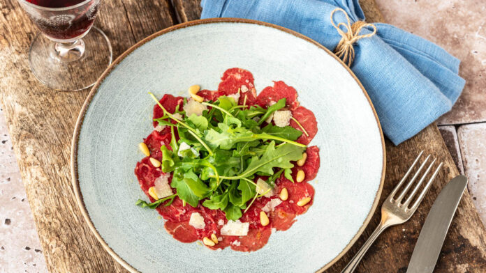 klassisches-carpaccio-vom-rind Ein Teller Carpaccio steht mit Wein auf Holzuntergrund - Foodblog Mundgefühl
