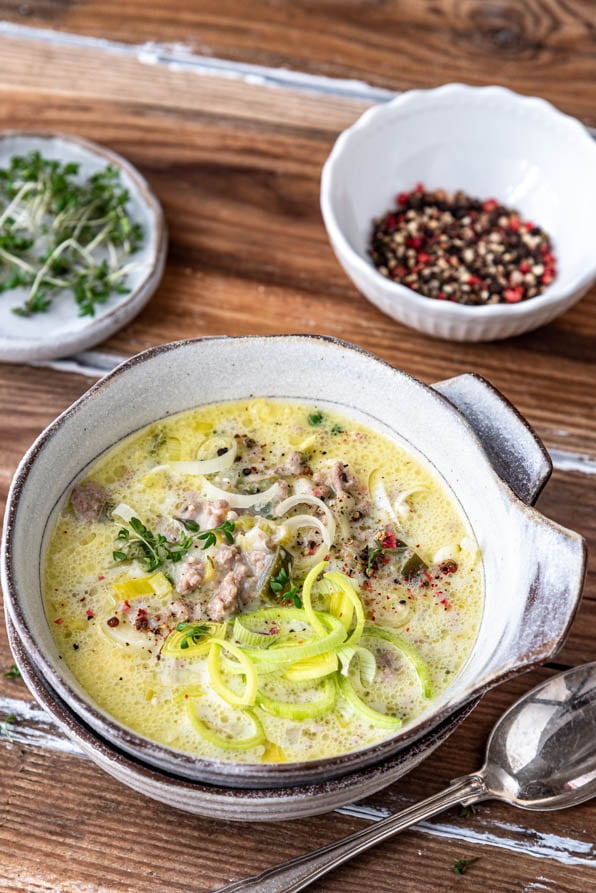 Käse-Lauch-Suppe in einem Schüsselchen auf Holzuntergrund. Daneben Pfeffer und Kresse.