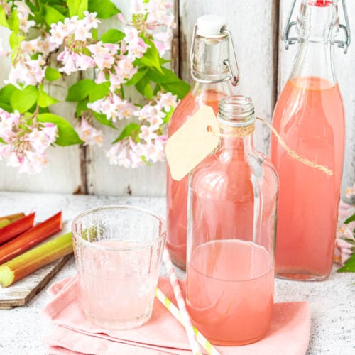 3 Flaschen Rhabarbersirup auf Holzuntergrund mit Rhabarberstangen