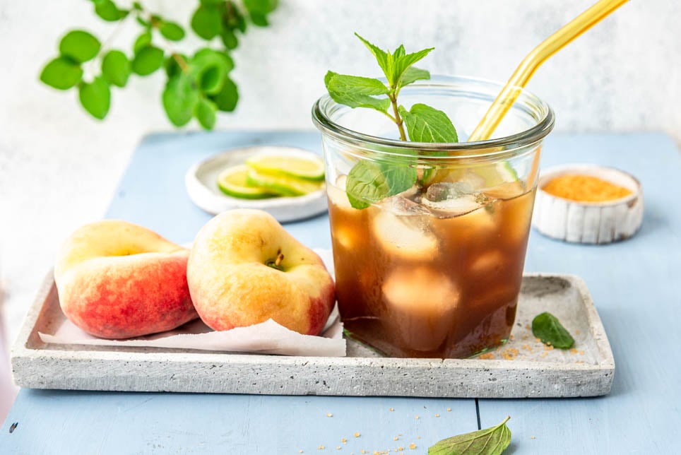 Glas mit selbstgemachtem Pfirsich Eistee, Pfirsichen und Eiswürfeln auf einem Tablett