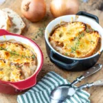 Goldbraun überbackene Französische Zwiebelsuppe mit Baguettescheiben und Kräutern in einer zwei kleinen Gratinformen serviert