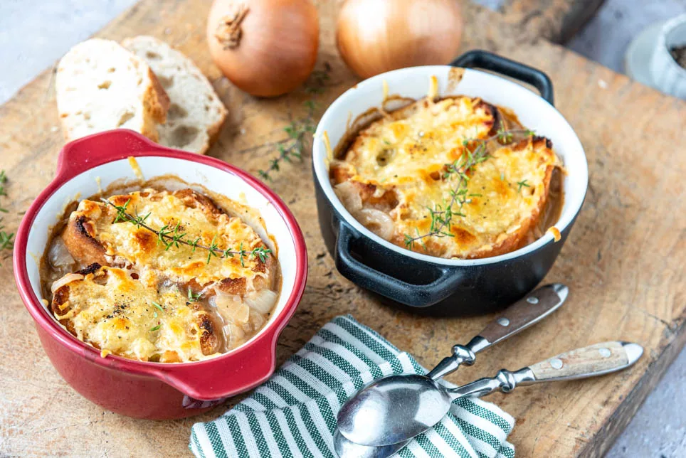 Goldbraun überbackene Französische Zwiebelsuppe mit Baguettescheiben und Kräutern in einer zwei kleinen Gratinformen serviert