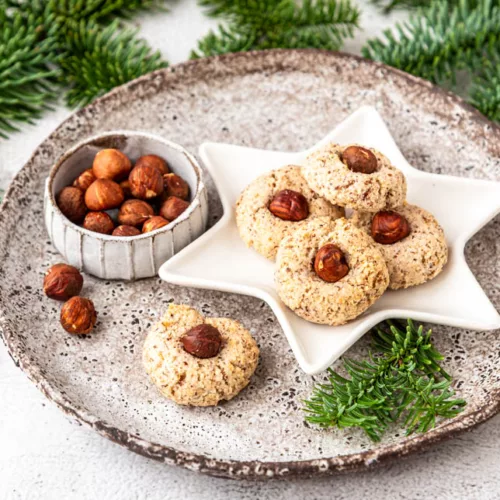 Saftige Haselnussmakronen mit ganzen Haselnüssen auf einem Sternenteller, angerichtet auf rustikalem Keramikteller mit Tannenzweigen im Hintergrund