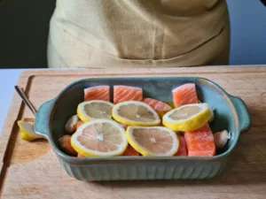 Lachs in einer Auflaufform auf einem Holzbrett, mit Zitronenscheiben und frischen Kräutern